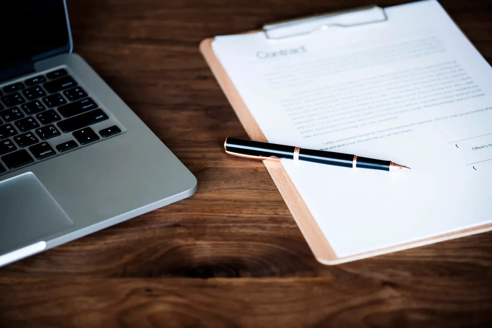 Contract on a clipboard with a pen, next to a laptop on a wooden desk